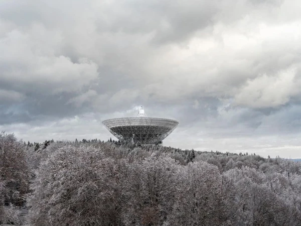 Wide shot of large radio telescope on a hill surrounded by snowy trees under a cloudy sky, reflecting cold, quiet technology and nature.