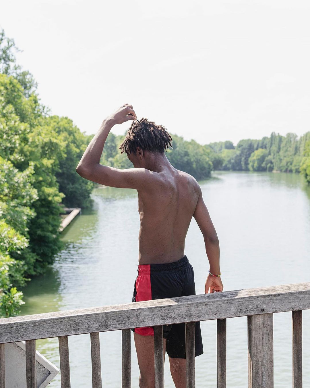 A shirtless African-American man with dreadlocks stands on a bridge, gazing at a river surrounded by green trees on a sunny day.