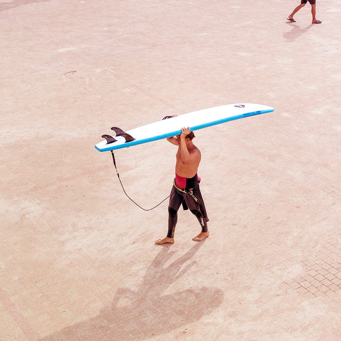Bare-chested adult male walking with surfboard overhead on tan surface, viewed from above with long shadow and distant figure.