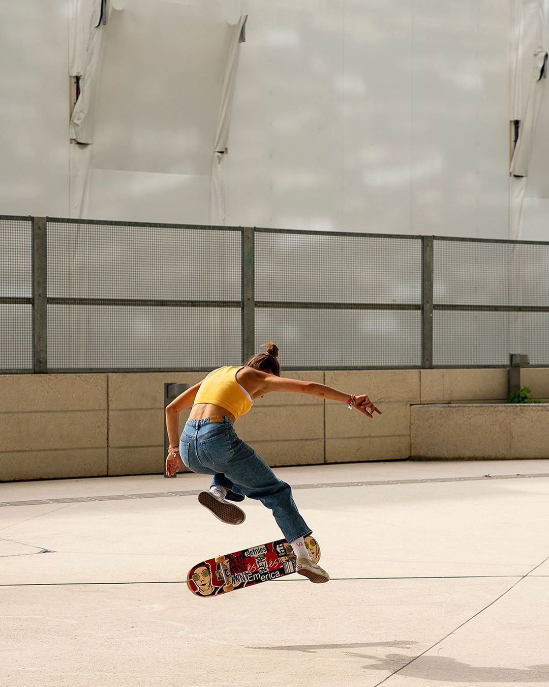 Female skateboarder performs mid-air trick on city pavement. Young woman in jeans, yellow tank top, and sneakers. Urban backdrop and daylight.
