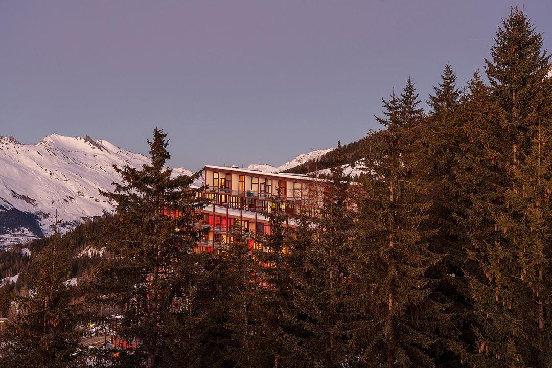 View of a modern building nestled among evergreen trees, with snow-covered mountains in the background under a blue sky.