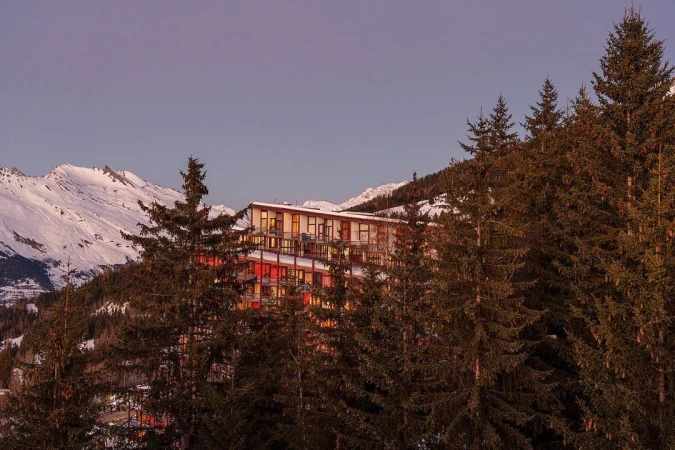 View of a modern building nestled among evergreen trees, with snow-covered mountains in the background under a blue sky.