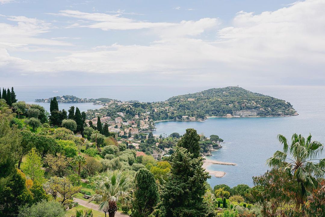 Coastal view featuring trees, buildings, and mountains along the French Riviera on a bright, sunny day with fluffy white clouds.