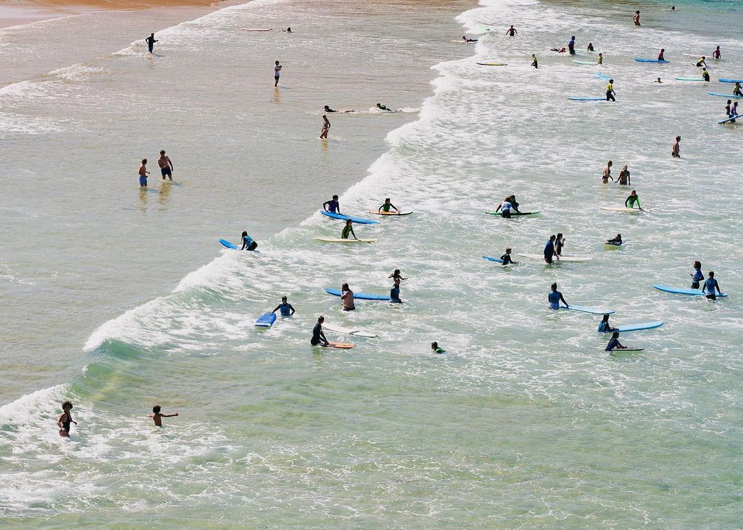 Aerial view of a crowded beach with many people swimming, surfing, and wading in the waves on surfboards in the ocean on a sunny day.