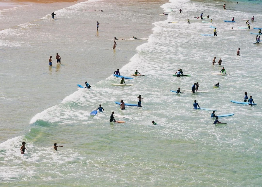 Aerial view of a crowded beach with many people swimming, surfing, and wading in the waves on surfboards in the ocean on a sunny day.