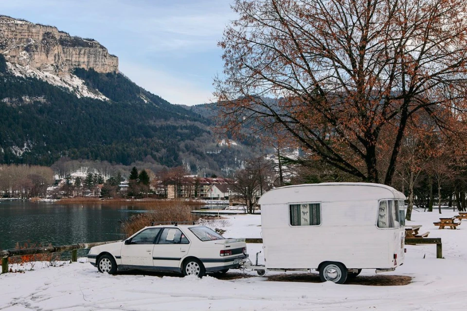White car towing a white caravan in a snowy landscape with a lake, mountains, and a tree with bare branches in the background.