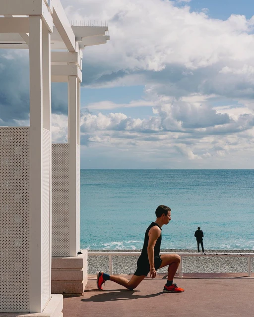 A man in athletic wear performs a lunge on a patio overlooking the ocean with a figure in the background and a white pergola.