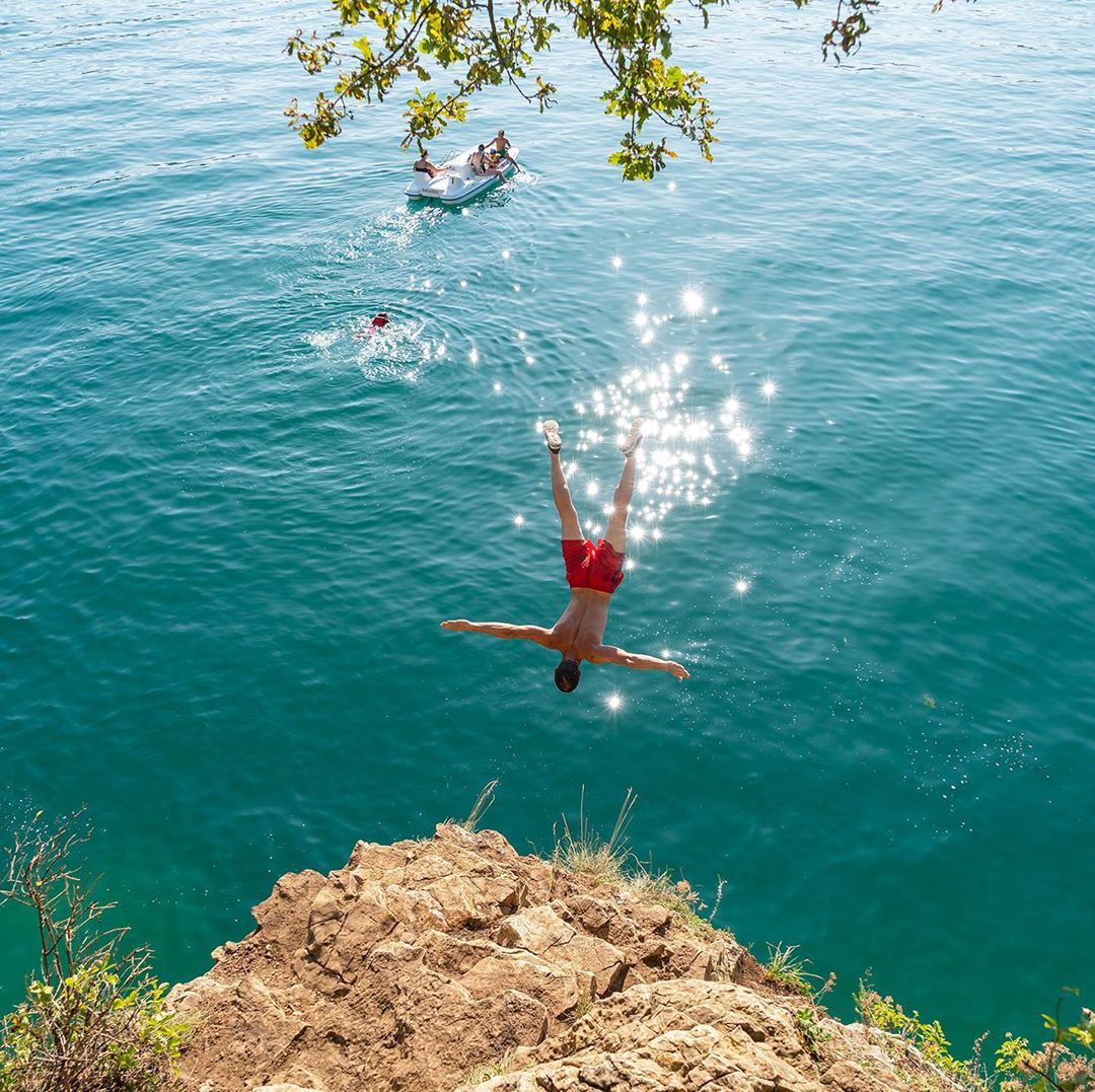 Adult male dives headfirst into turquoise water off a rocky cliff, bathed in sunlight. People on a raft in the background.