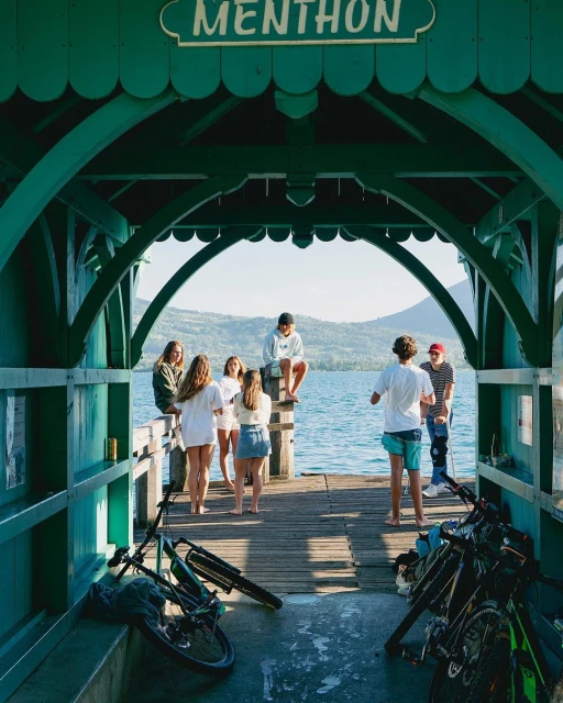 Group of young adults gather on a wooden pier under a green archway with bicycles, overlooking a calm lake and distant mountains.