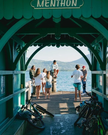 Group of young adults gather on a wooden pier under a green archway with bicycles, overlooking a calm lake and distant mountains.