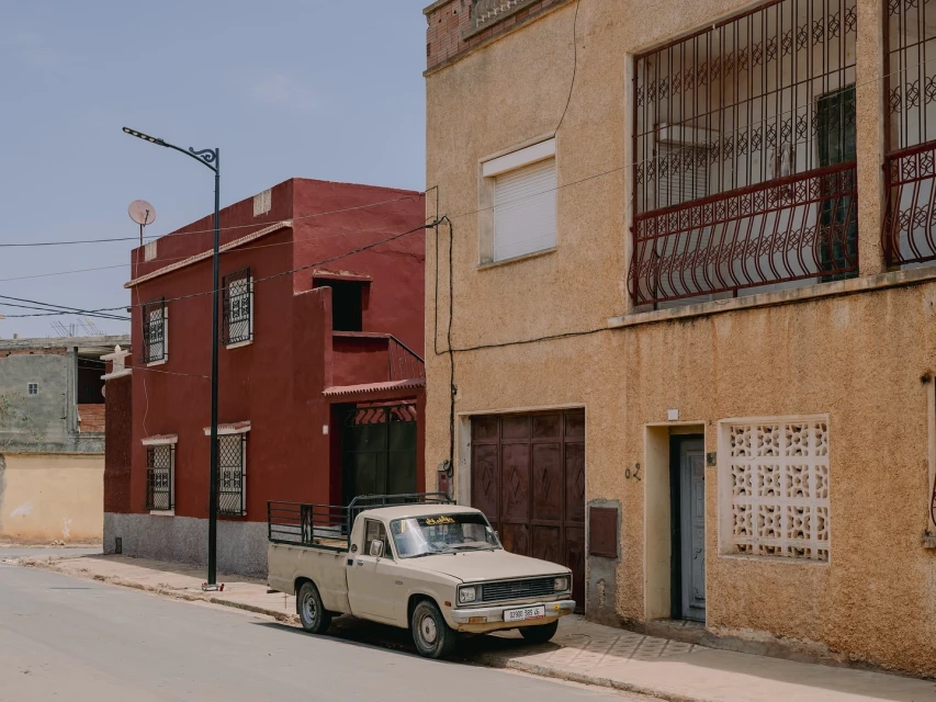 Beige pickup truck parked on a street next to a red building and a beige building with a blue sky overhead in an outdoor daytime shot.