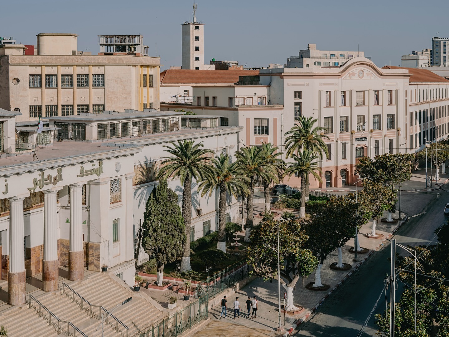 Elevated view of colonial architecture, palm trees, and city street with a few people walking on the sidewalk in Constantine, Algeria.