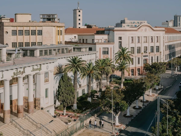 Elevated view of colonial architecture, palm trees, and city street with a few people walking on the sidewalk in Constantine, Algeria.