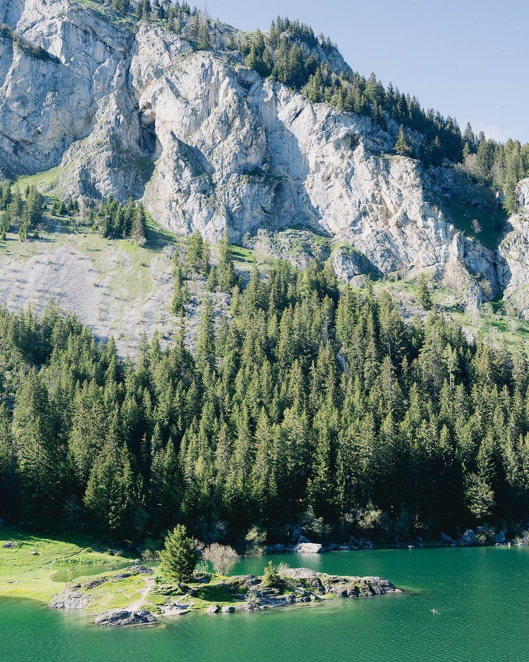 Scenic landscape featuring a mountain, forest, and lake. The rugged mountain face rises above a dense green forest and turquoise lake.