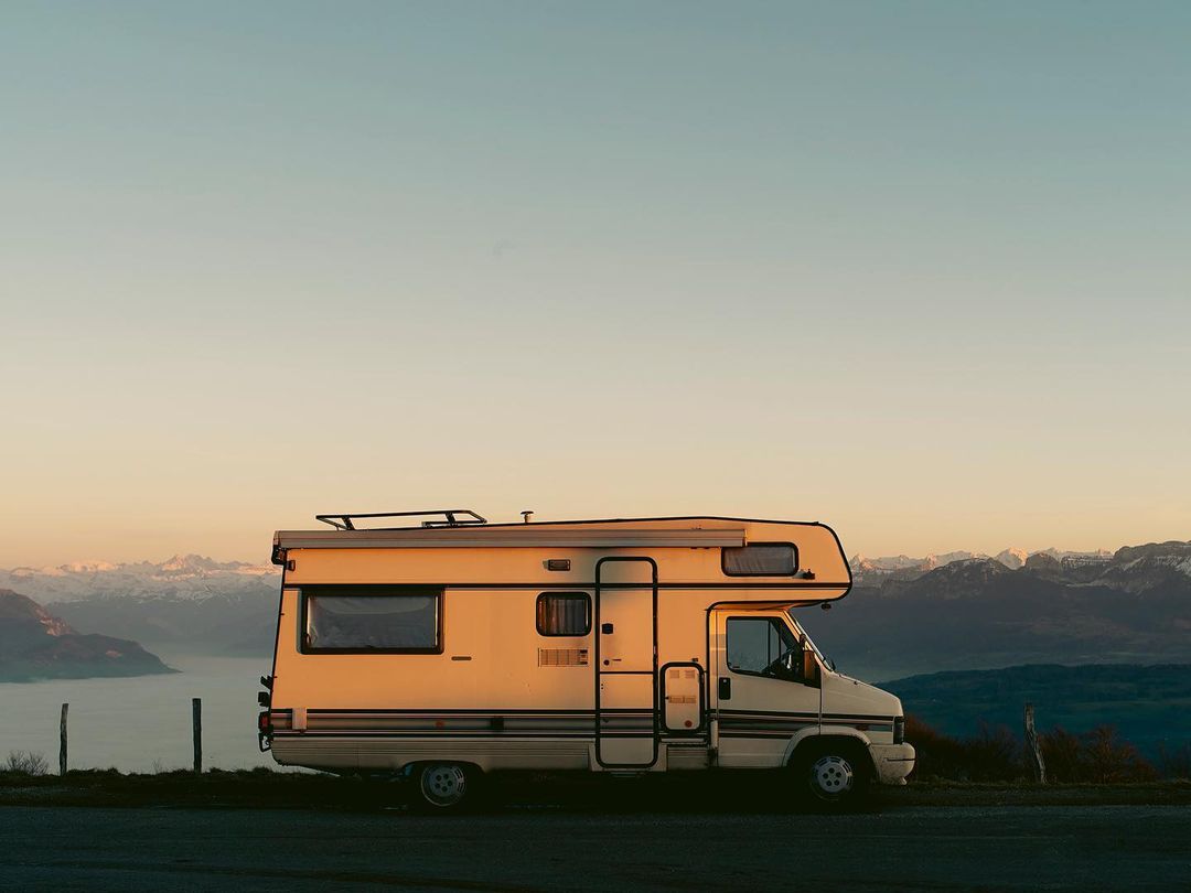 Beige camper van parked on a road with a mountain and lake landscape in the background during sunset, casting a warm glow.