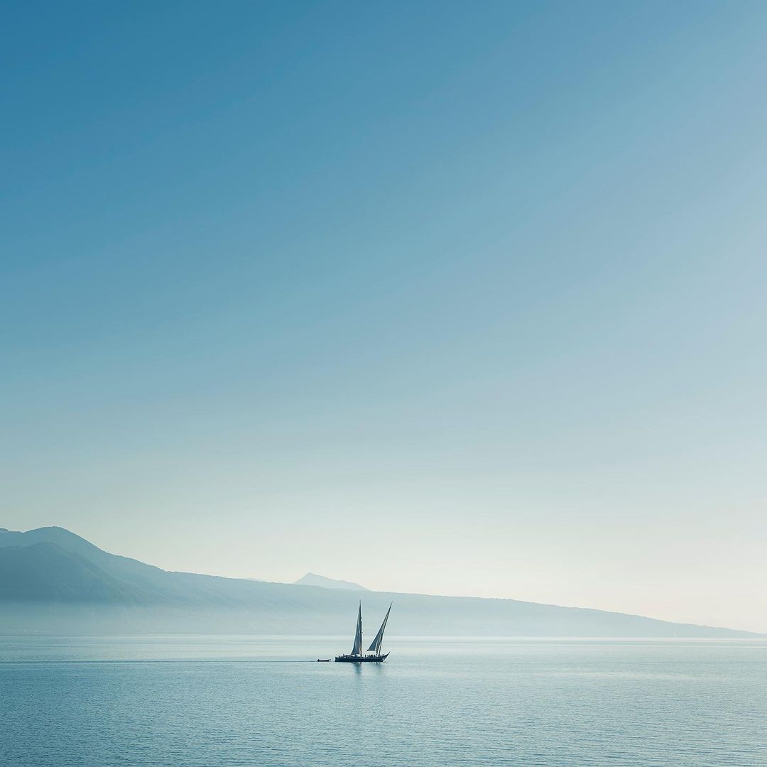 Sailboat glides on the water under a clear sky with distant mountains in the background, creating a serene and minimalist marine vista.