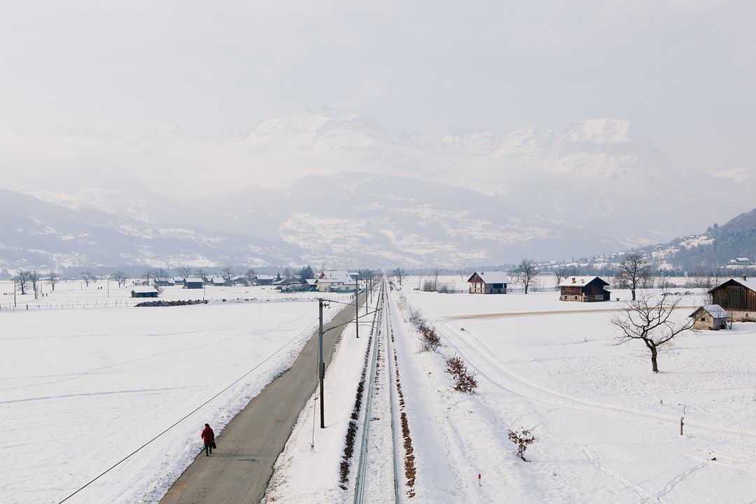 Winter landscape with snow-covered fields, village houses, mountains, and a person in red coat walking along road near train tracks.