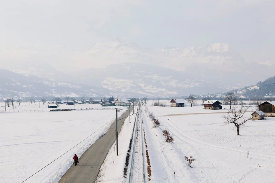 Winter landscape with snow-covered fields, village houses, mountains, and a person in red coat walking along road near train tracks.
