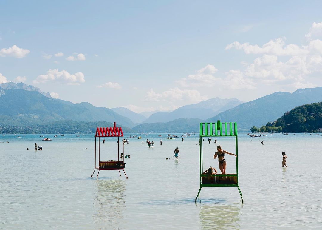 People enjoy leisure time at the beach with shallow, clear water, mountains in the background, and framed by colorful artistic lifeguard stands.