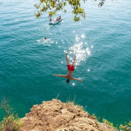 Man diving headfirst from a rocky cliff into clear turquoise water with a small white boat visible in the distance