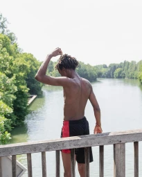A shirtless young Black man standing on a wooden bridge, adjusting his dreadlocks with a river and trees in the background.