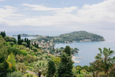 High-angle view overlooking a Mediterranean coastal town, lush green hills, and blue sea under a partly cloudy sky.