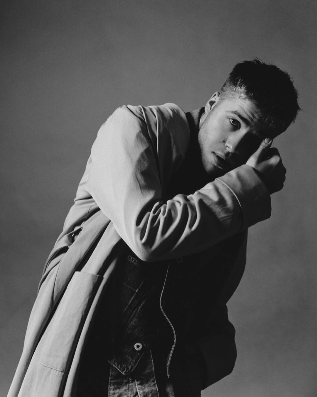 Black and white portrait of a man in a coat with hand near his head against a neutral grey studio background