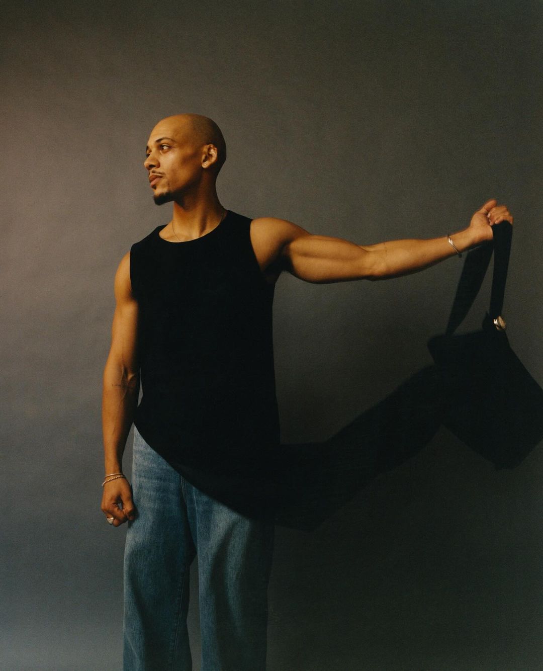 Man in black tank top and jeans poses with a black bag against a gray background in a studio shot with soft lighting.