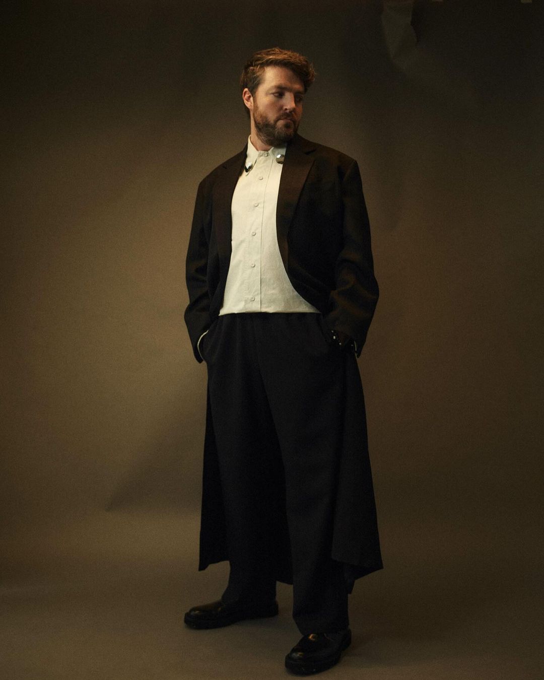Standing man in a formal black tuxedo and trousers with a light green shirt, posing in a studio setting with warm, diffused lighting.