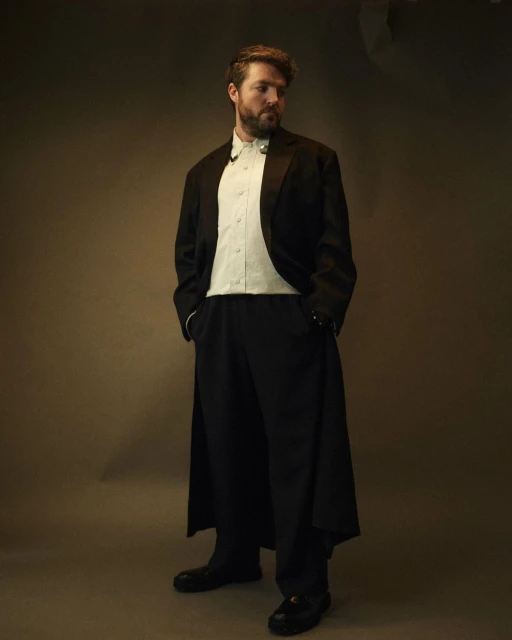 Standing man in a formal black tuxedo and trousers with a light green shirt, posing in a studio setting with warm, diffused lighting.