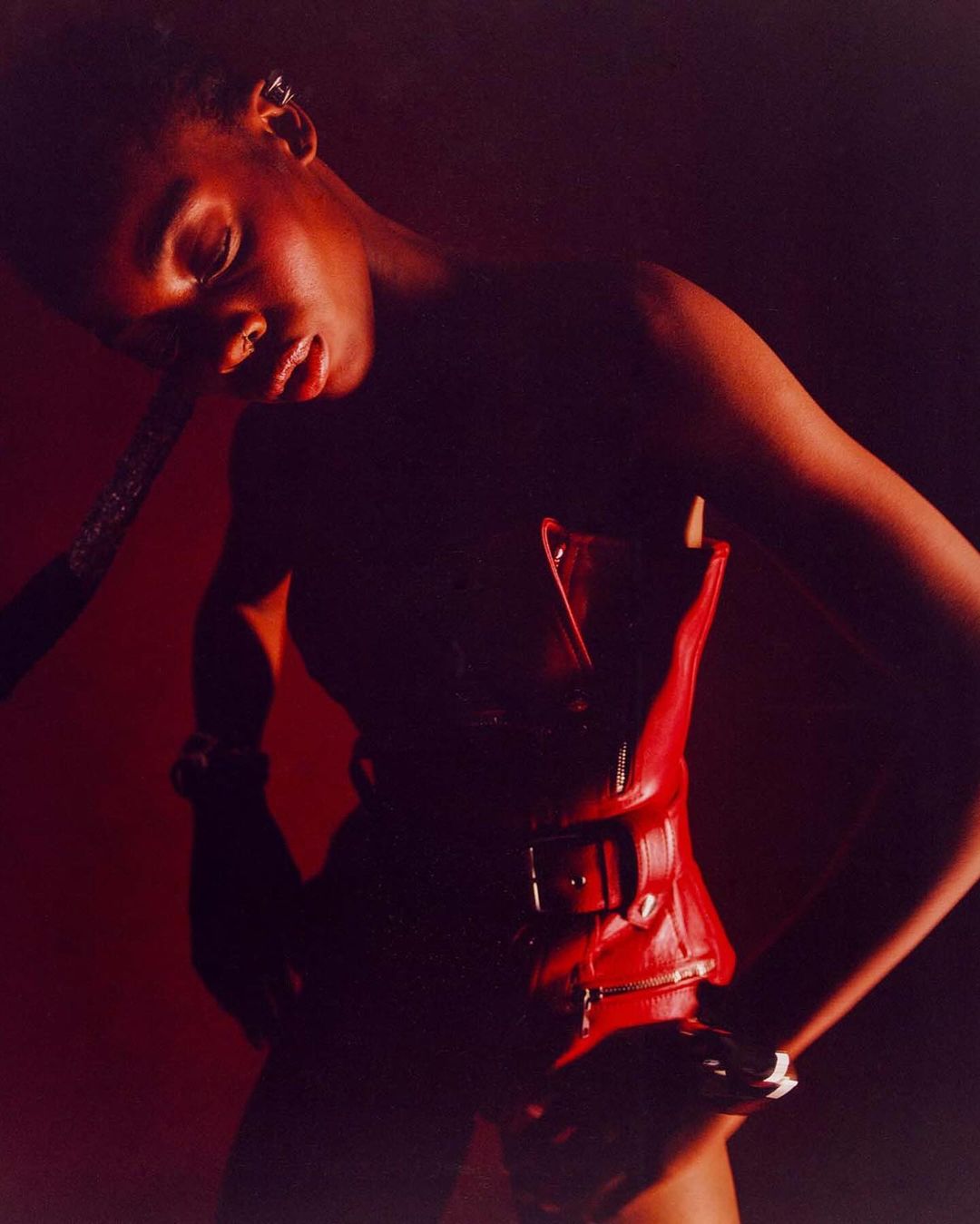 A Black female model strikes a pose in a close-up studio shot, wearing a red leather corset and dark hair against a deep red backdrop.