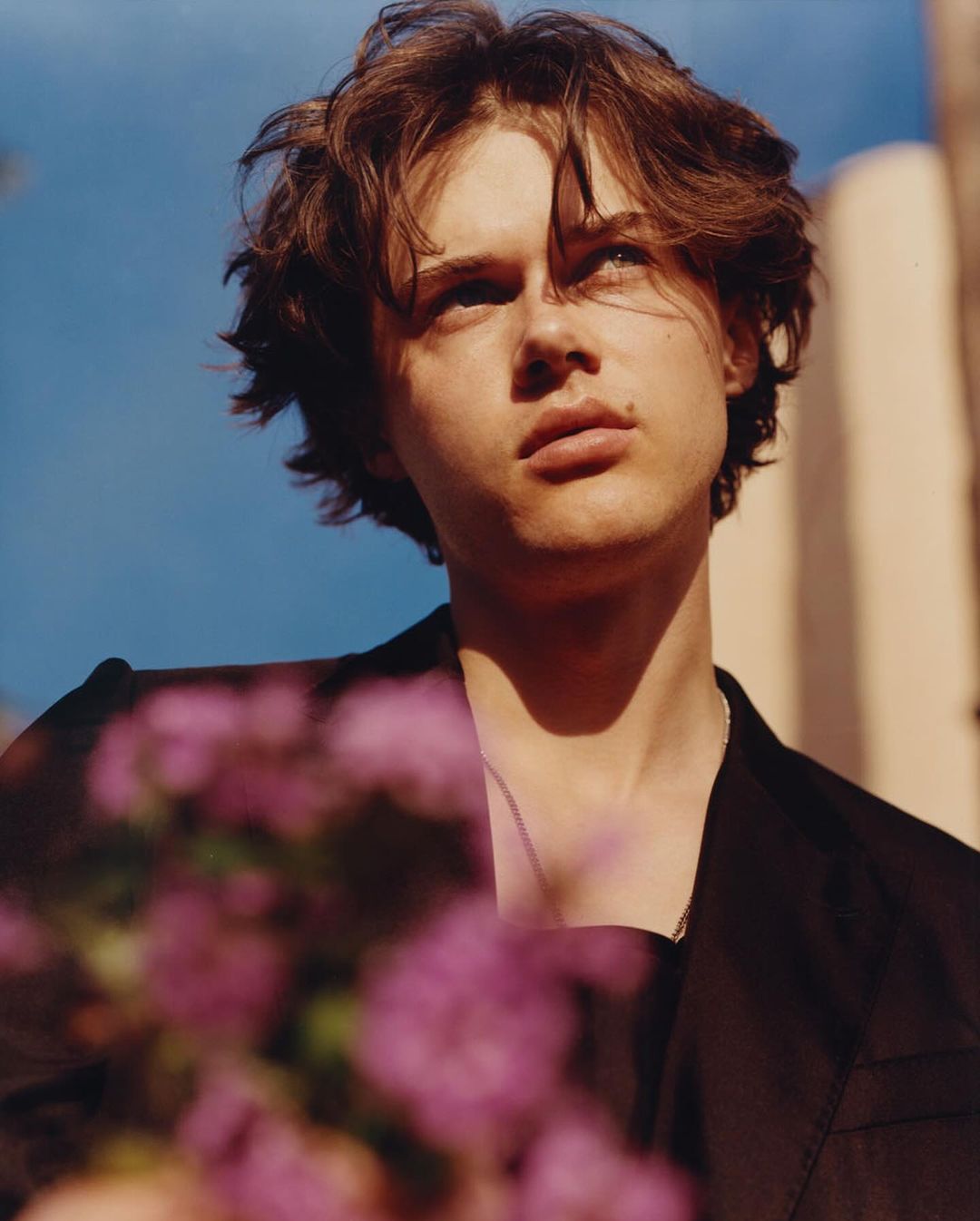 Low-angle portrait of a young man with medium-length brown hair wearing a black suit jacket with purple flowers in the foreground against blue sky.