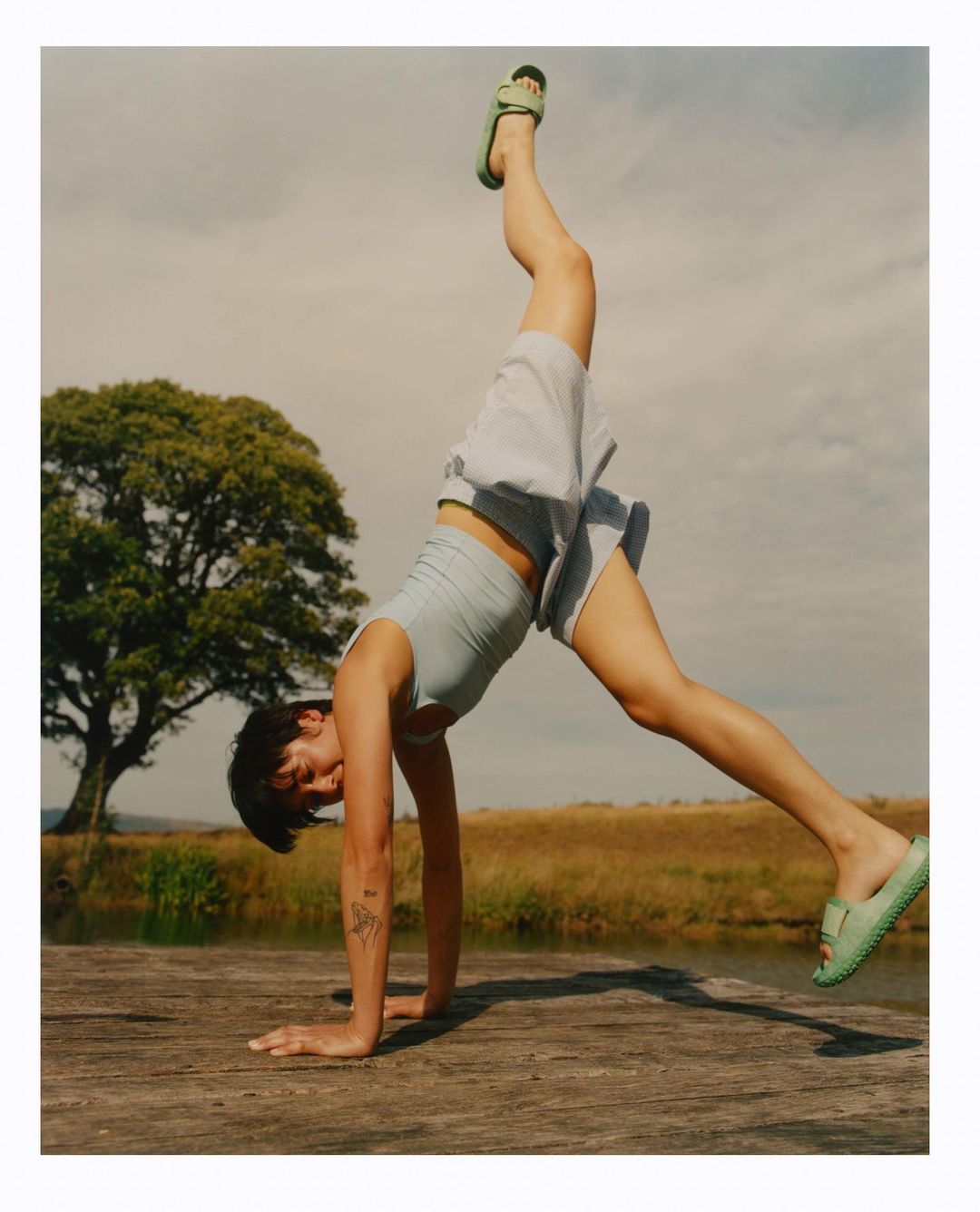 Young woman in sportswear does a handstand on a wooden deck with a tree, field, and cloudy sky in the background on a sunny day.