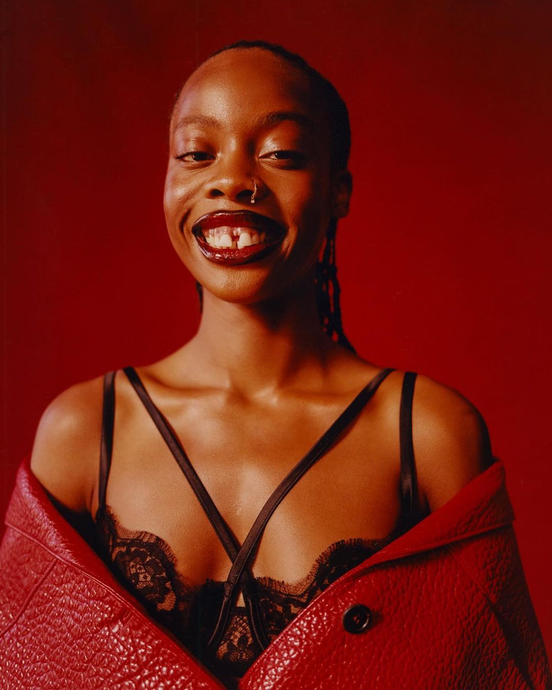 Close-up studio portrait of an African woman smiling, wearing a black lace bra, red jacket, and dark lipstick against a red background.