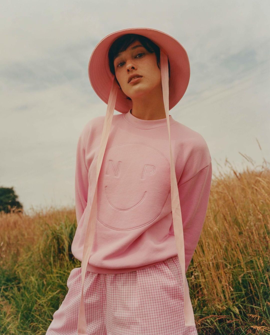 A model in a light pink hat and matching outfit poses in a field of tall grass under a cloudy sky, creating a soft, dreamy aesthetic.