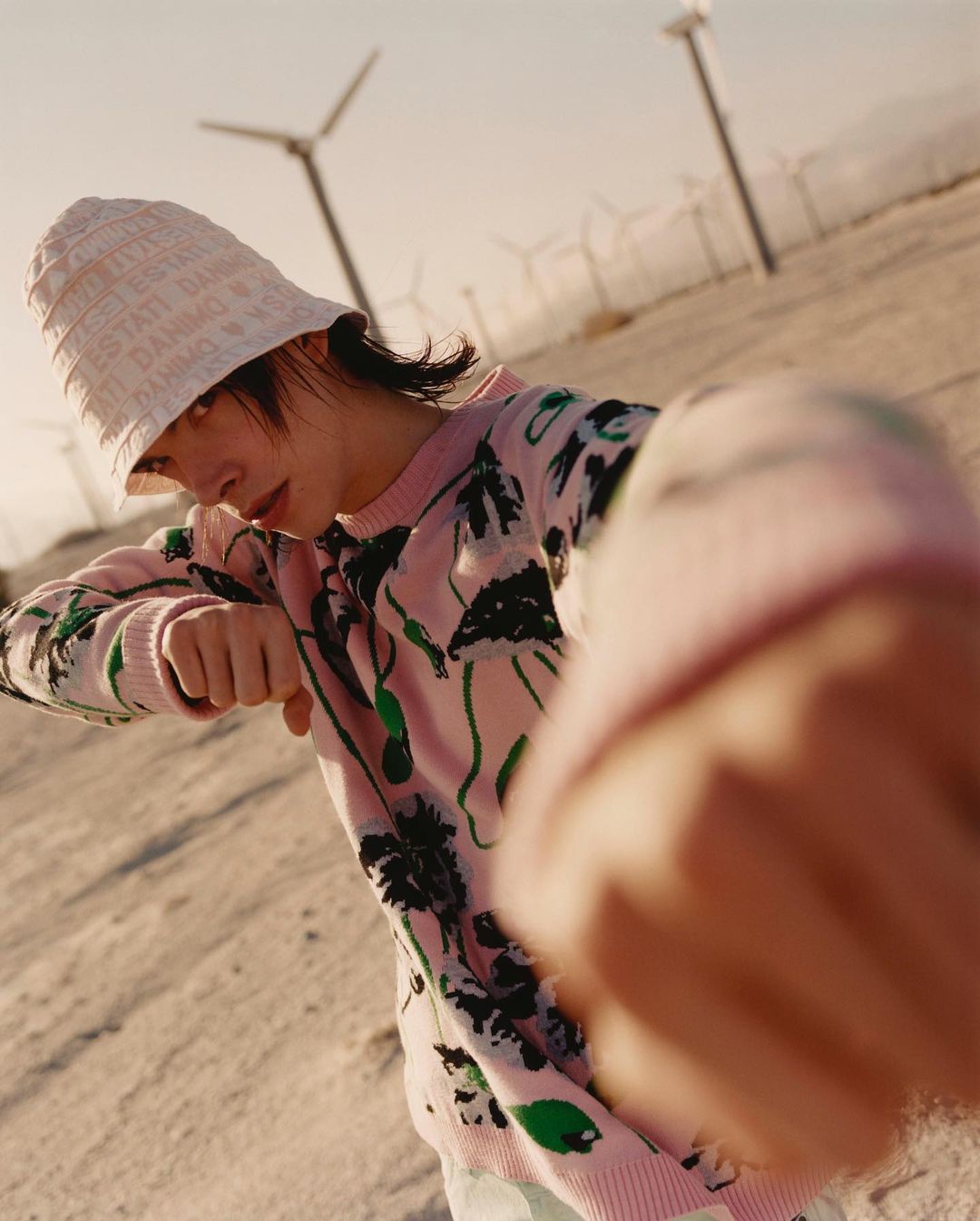 Eye-level shot of a person in a patterned pink sweater and bucket hat standing in a desert landscape with wind turbines.