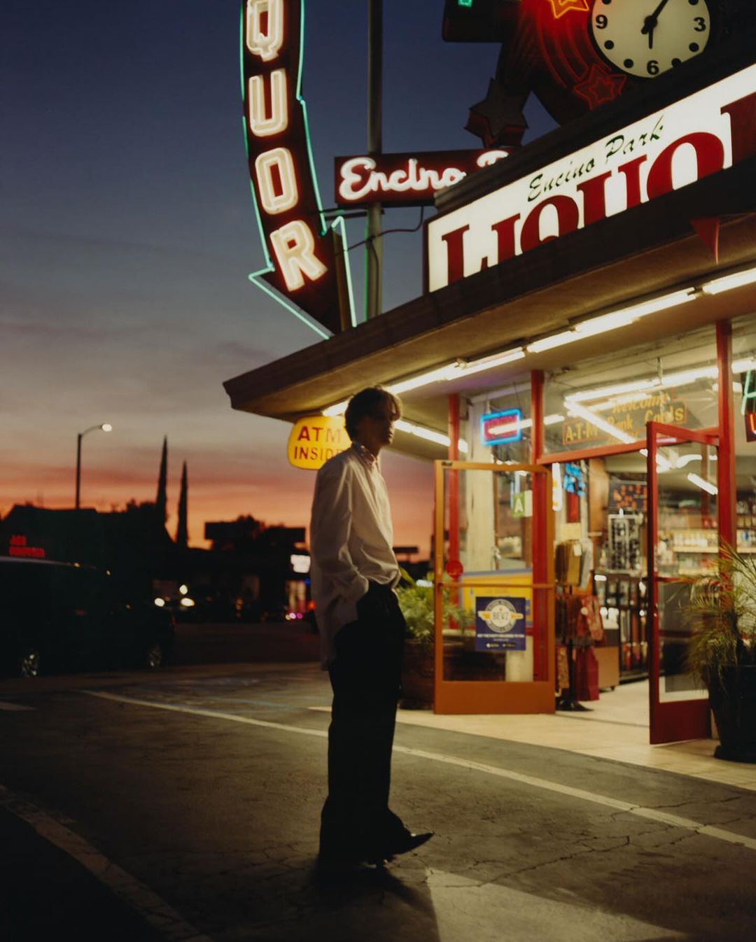 Man stands outside a liquor store at dusk; the store's neon lights glow against the twilight sky, creating a vintage, cinematic feel.