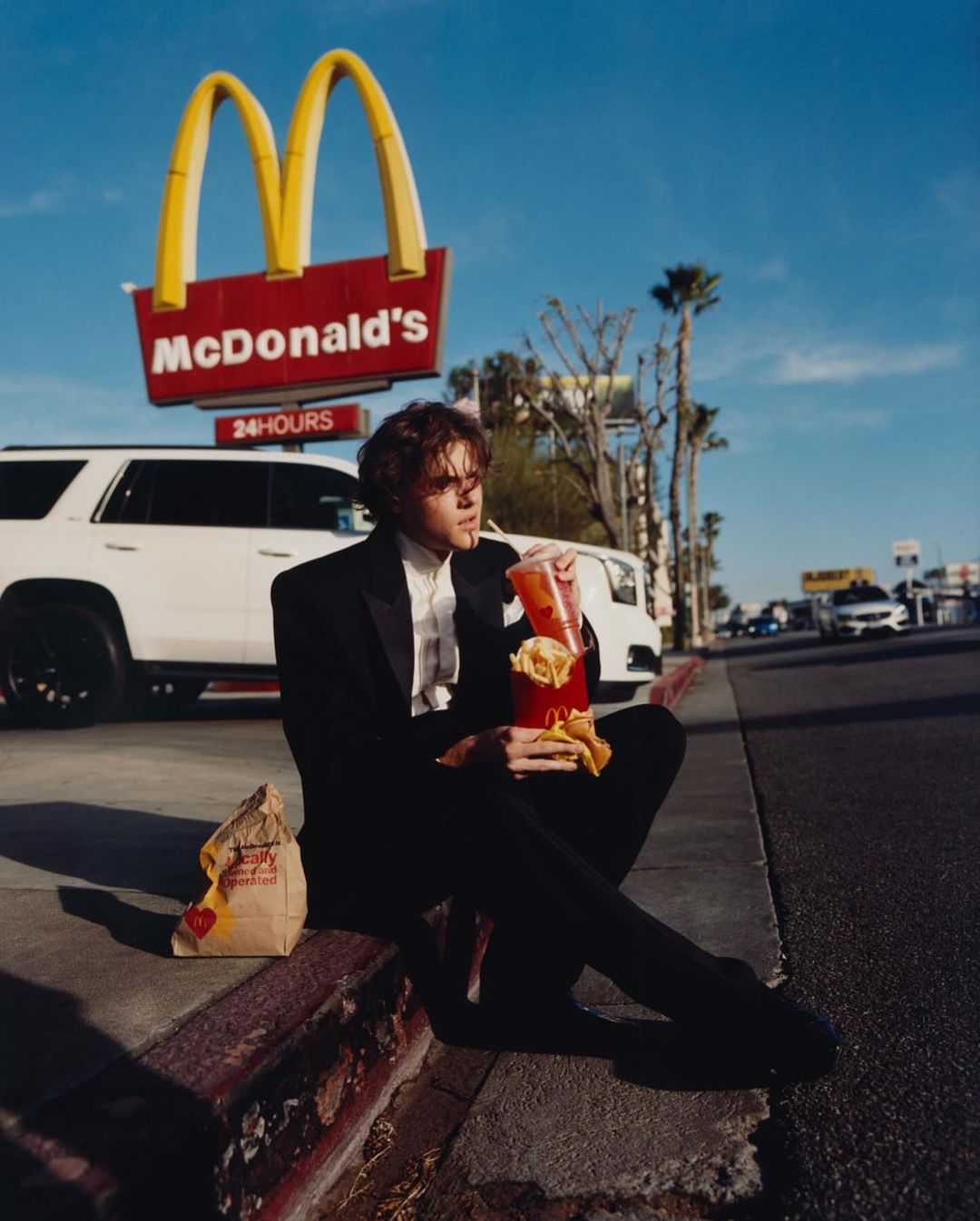 Stylish adult male in black suit eats fast food on sidewalk near McDonald's sign, with passing cars and palm trees in the background.