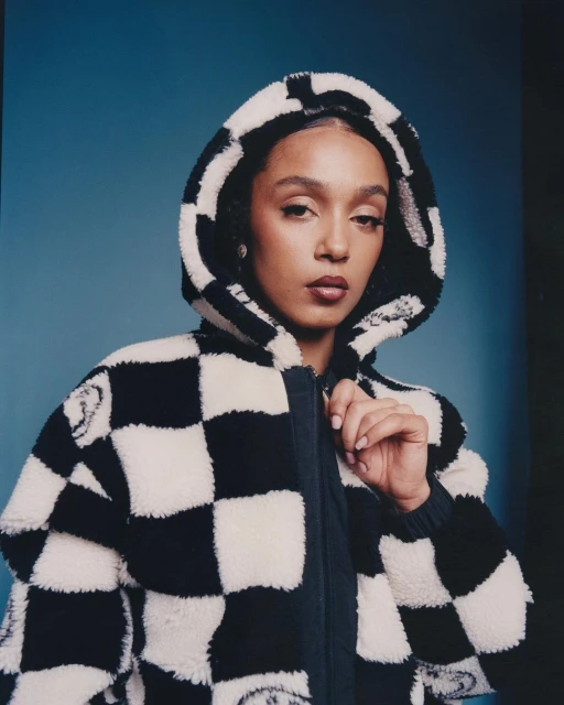 Portrait of a woman with dark hair wearing a black and white checkered jacket with a hood against a blue background.