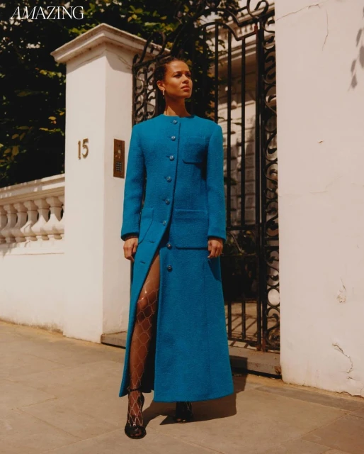 A stylish adult female in a long teal coat stands on a sidewalk near a gate, bathed in sunlight, for a fashion editorial shot outdoors.
