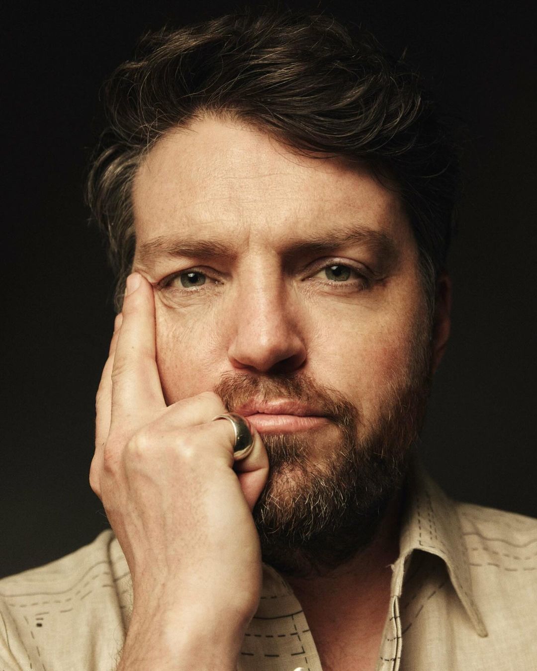 Headshot of an adult male with dark hair and a beard, wearing a patterned shirt, with his hand near his face, against a dark background.
