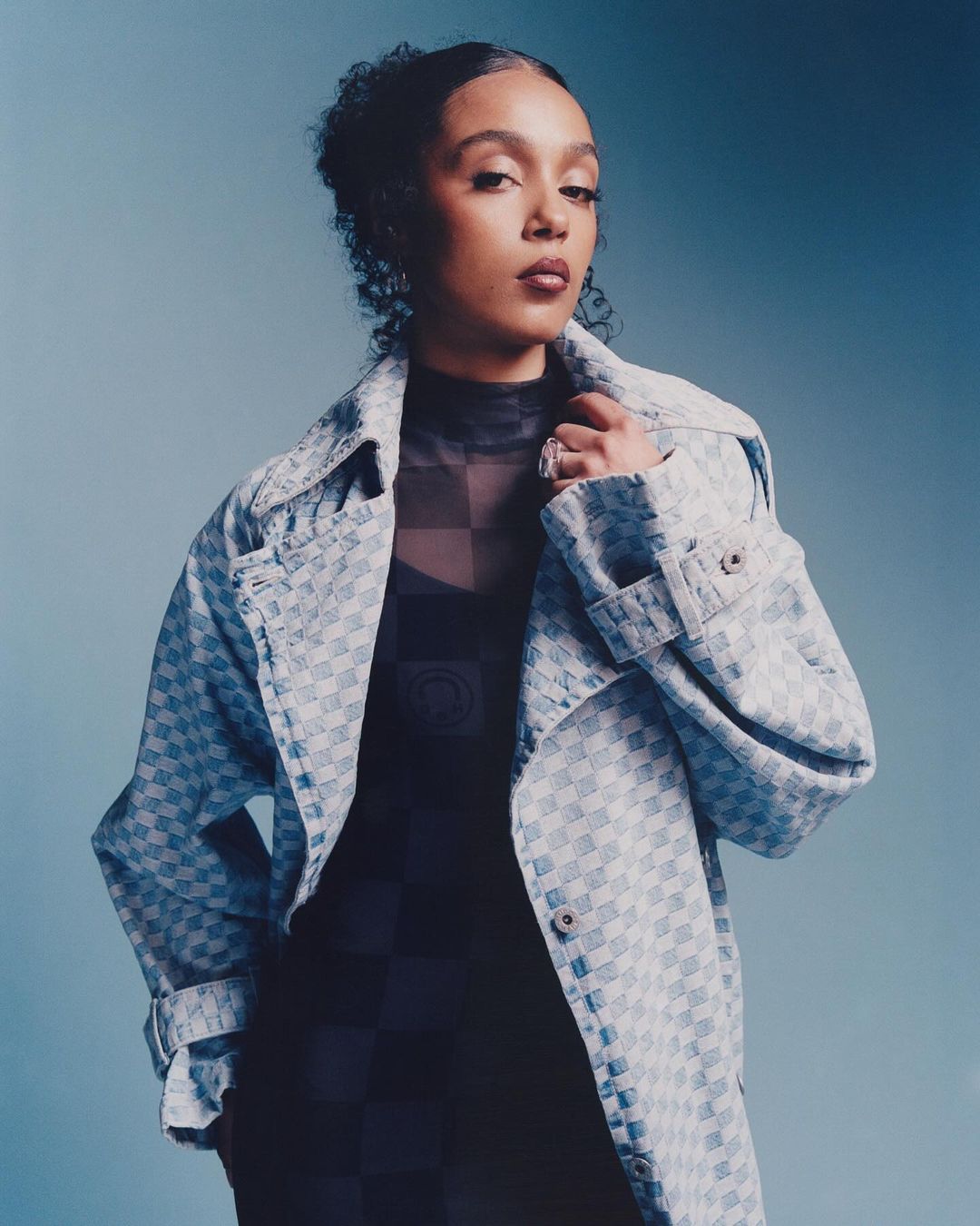 Portrait of a young woman with curly hair wearing a patterned blue coat and black checkered top against a gradient blue background.