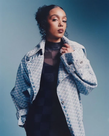 Portrait of a young woman with curly hair wearing a patterned blue coat and black checkered top against a gradient blue background.