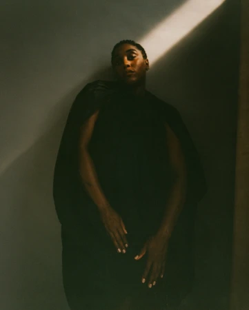 Dramatic portrait of a Black adult in dark clothing, captured in a photo studio with a single light source and a medium studio shot.