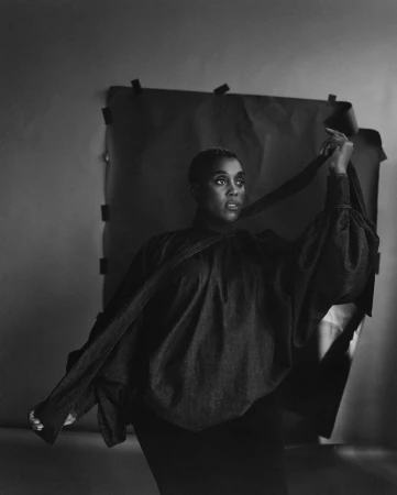 Portrait of a Black woman in a dark puffed-sleeve blouse holding fabric against a taped backdrop in a black and white photograph by Rhys Frampton.