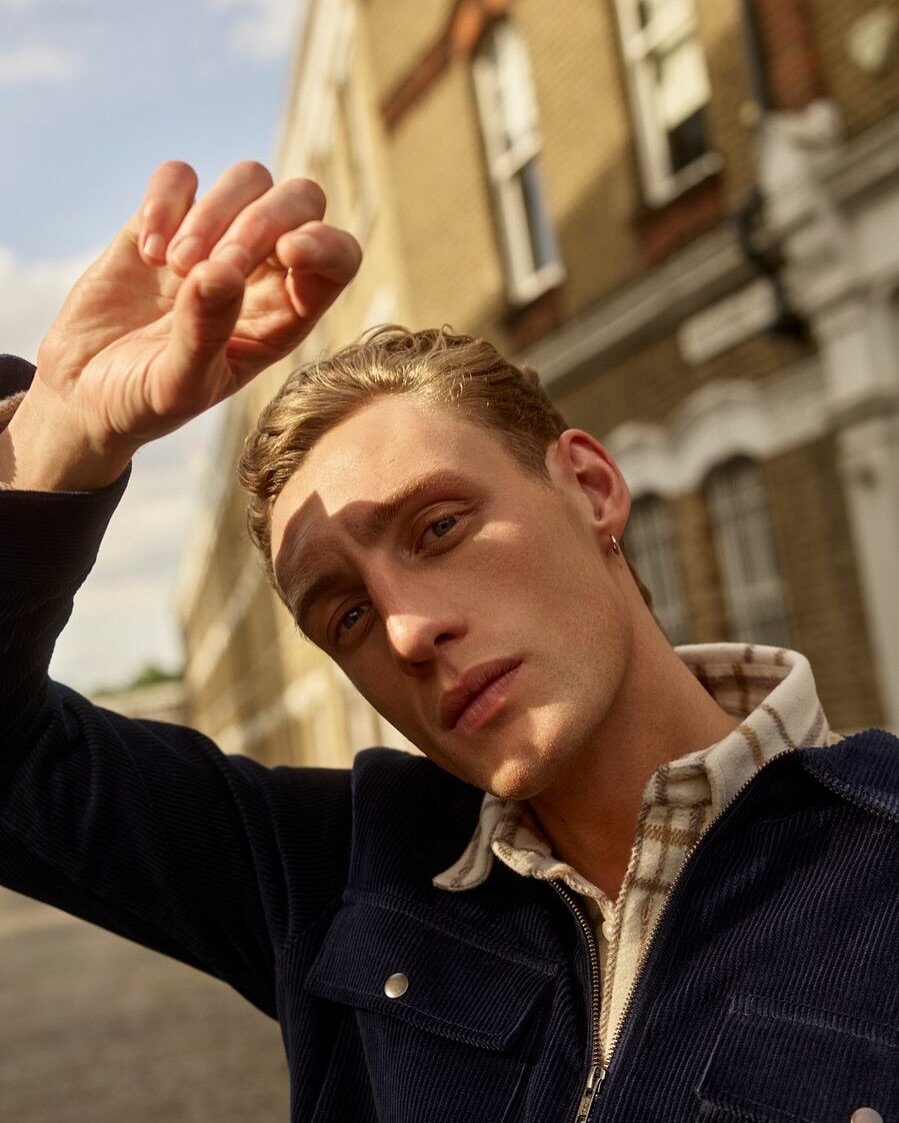 Portrait of a young adult man with blond hair, wearing a blue corduroy jacket and patterned shirt, posing outdoors against a brick building.