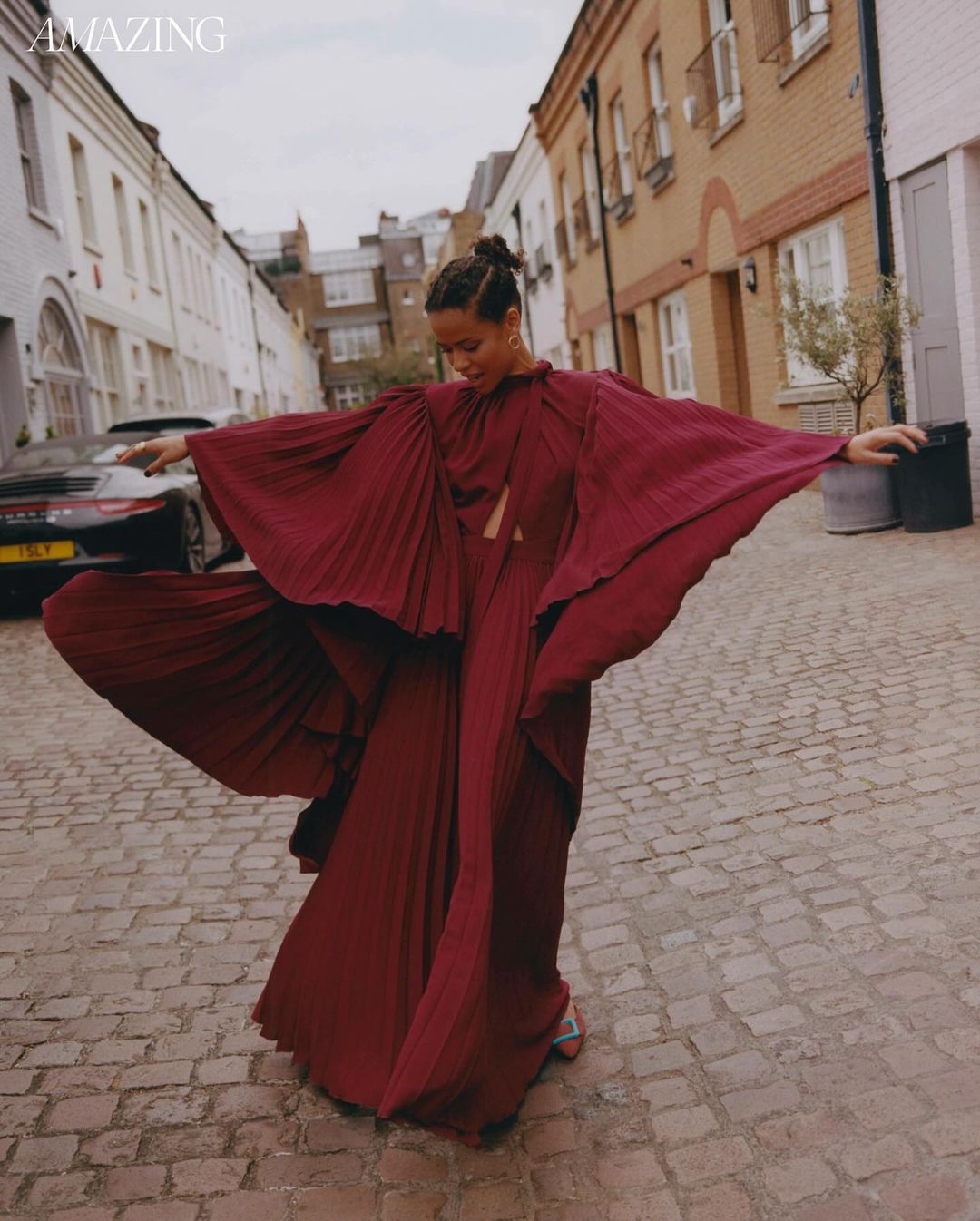 Woman in burgundy dress with outstretched arms on cobblestone street, brick buildings, daytime, overcast, low angle, fashion editorial.