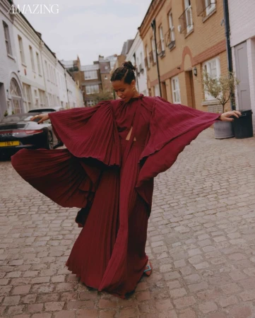 Woman in burgundy dress with outstretched arms on cobblestone street, brick buildings, daytime, overcast, low angle, fashion editorial.