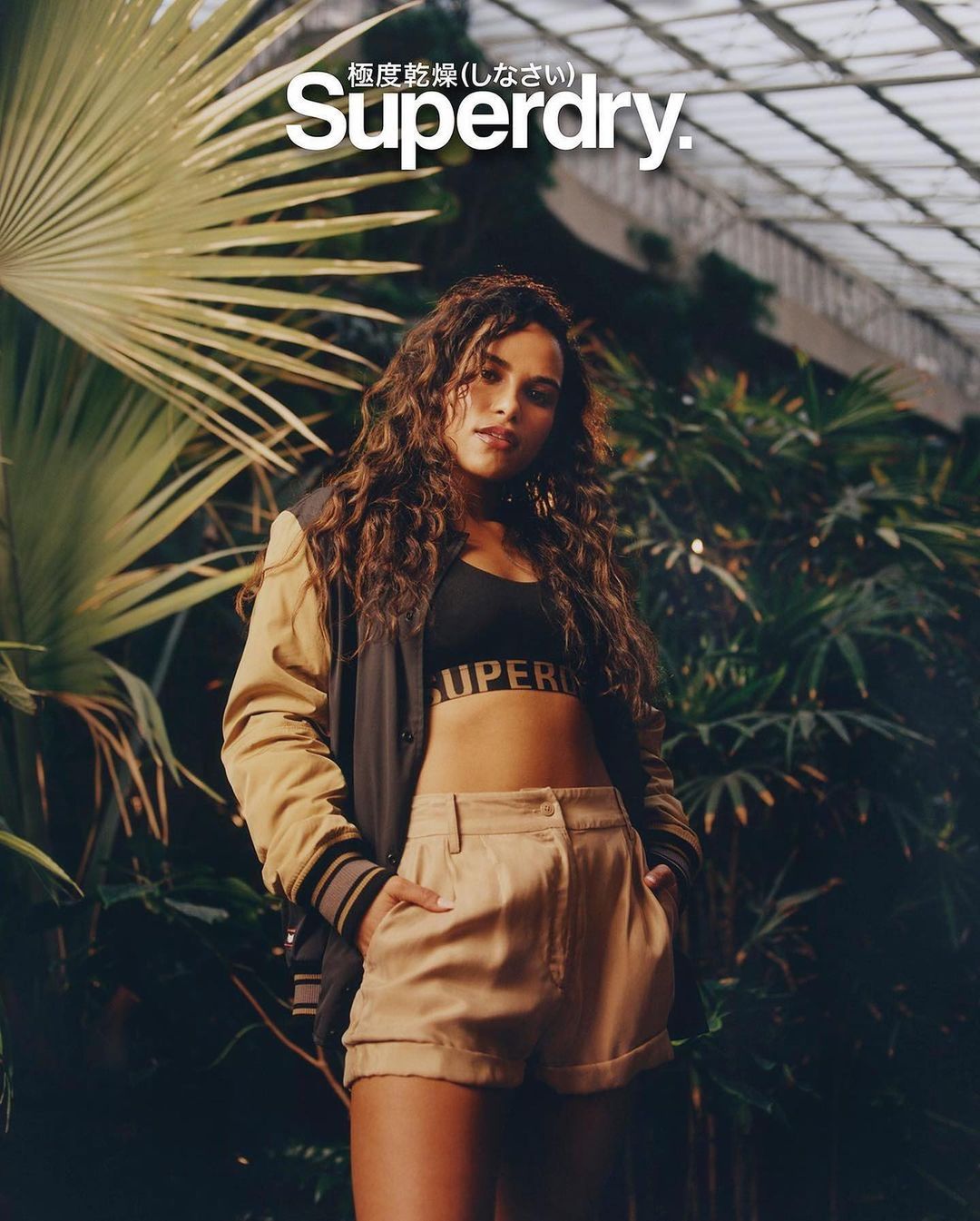 A young woman poses in athleisure wear, a cropped tank top and shorts, against a backdrop of dense greenery and a blurred glass ceiling.