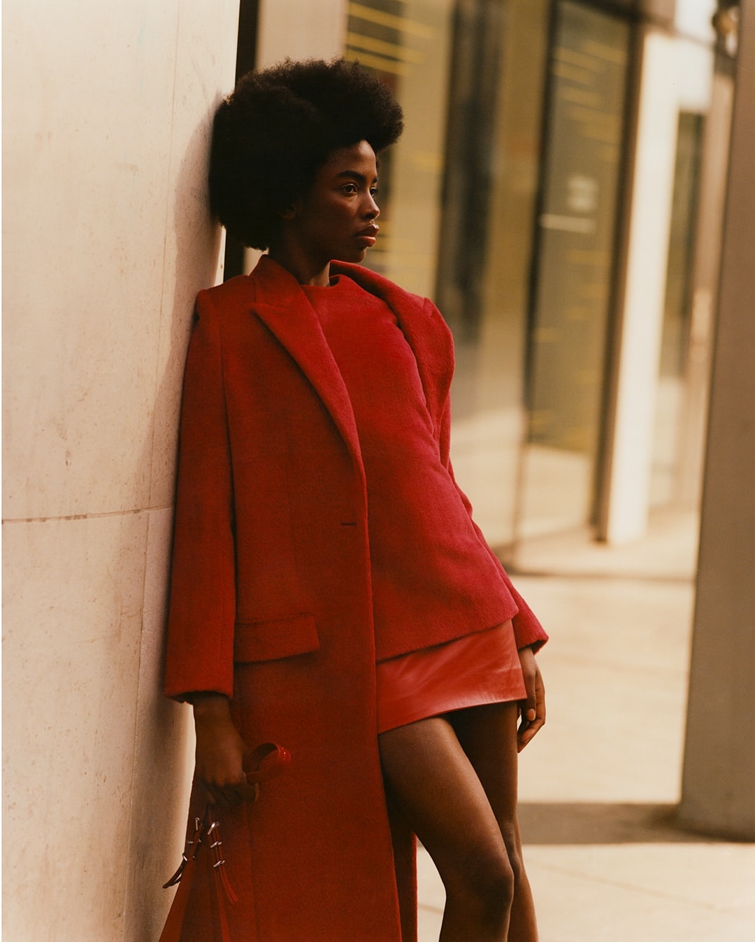 Stylish young Black woman in a red coat and skirt leans against an urban building, showcasing fashion in an outdoor setting.